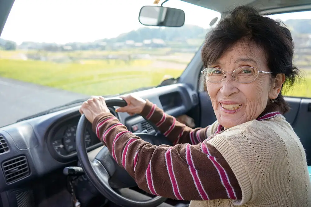 Image of older woman in driver seat of car. She has her hands on the steering wheel and is looking at camera over her shoulder